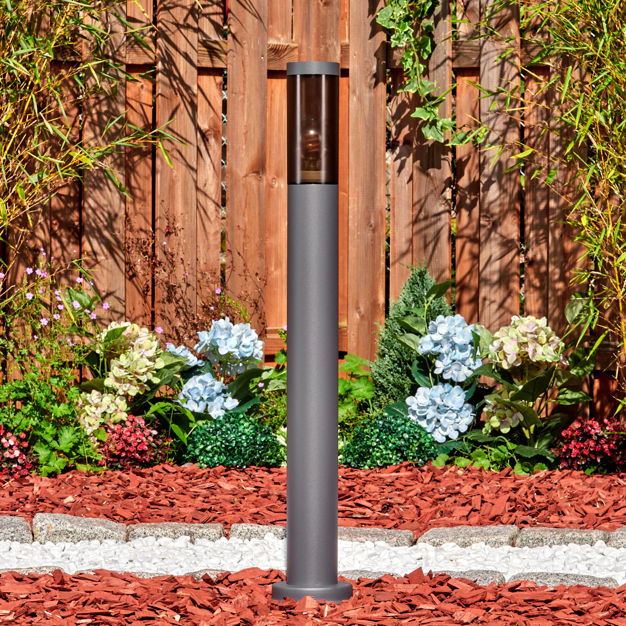 Eine graue Außenlampe steht in einem Garten mit roter Mulch, Blumen, einem weißen Steinrand und einem Holzzaun im Hintergrund.