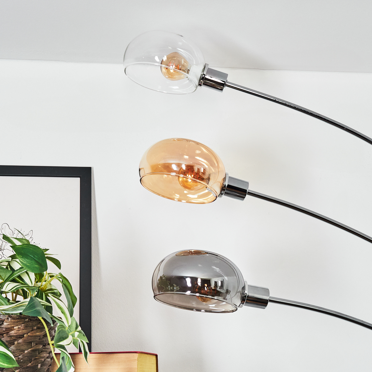 Eine moderne Stehlampe mit drei Glasschirmen. Einer ist klar, einer bernsteinfarben und einer verspiegelt. Eine Pflanze und gerahmte Kunst im Hintergrund.