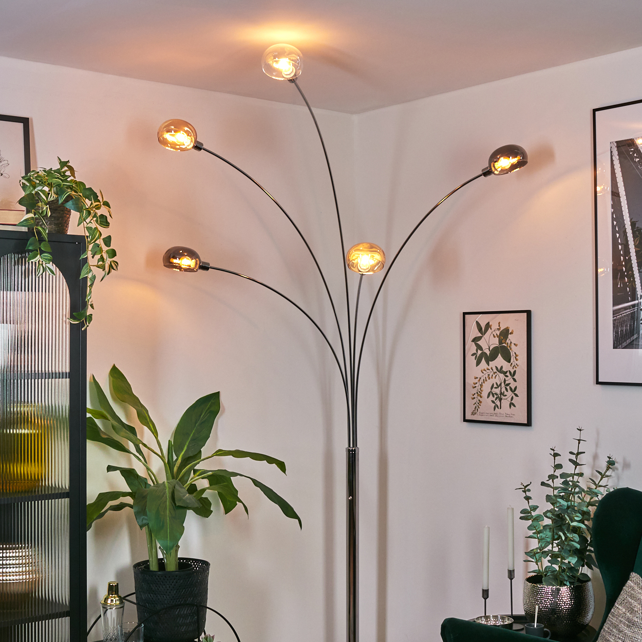Eine moderne Stehlampe mit fünf Glühbirnen beleuchtet einen Raum mit Pflanzen, einem schwarzen Schrank und gerahmten Kunstwerken. Die Szene ist gemütlich.