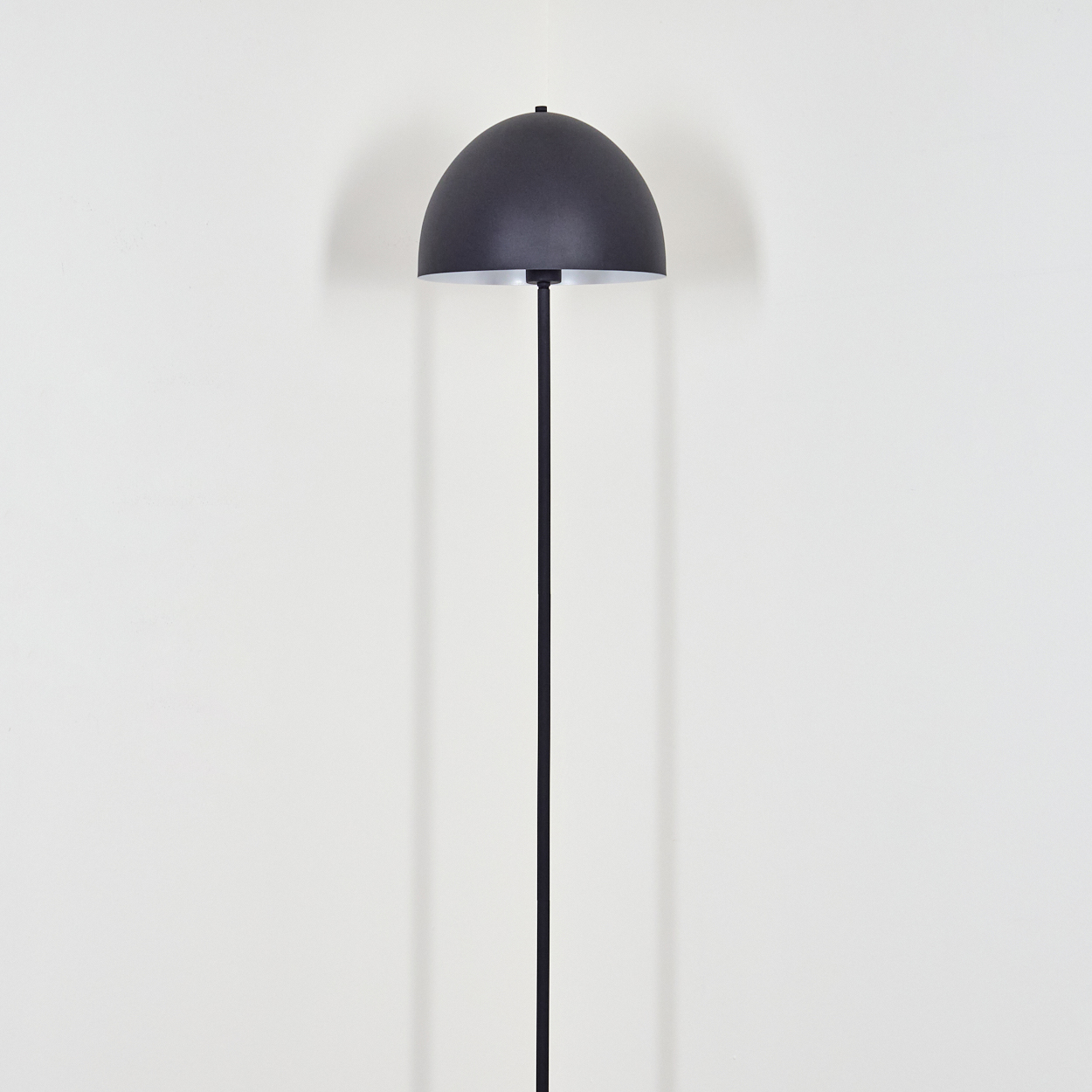 Eine moderne schwarze Stehlampe mit einem halbkugelförmigen Schirm steht vor einer weißen Wand. Die Lampe hat einen schlanken schwarzen Stiel.