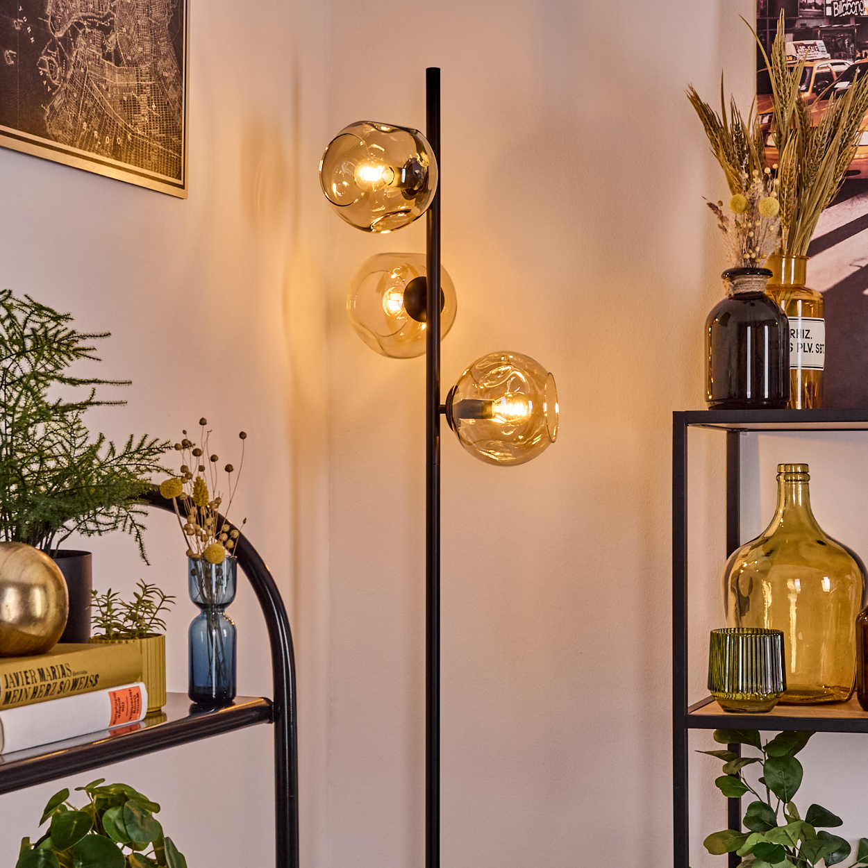 Eine moderne Stehlampe mit drei Kugelleuchten. Regale halten Vasen, Bücher und Pflanzen. Eine Karte und ein Poster hängen an der Wand.