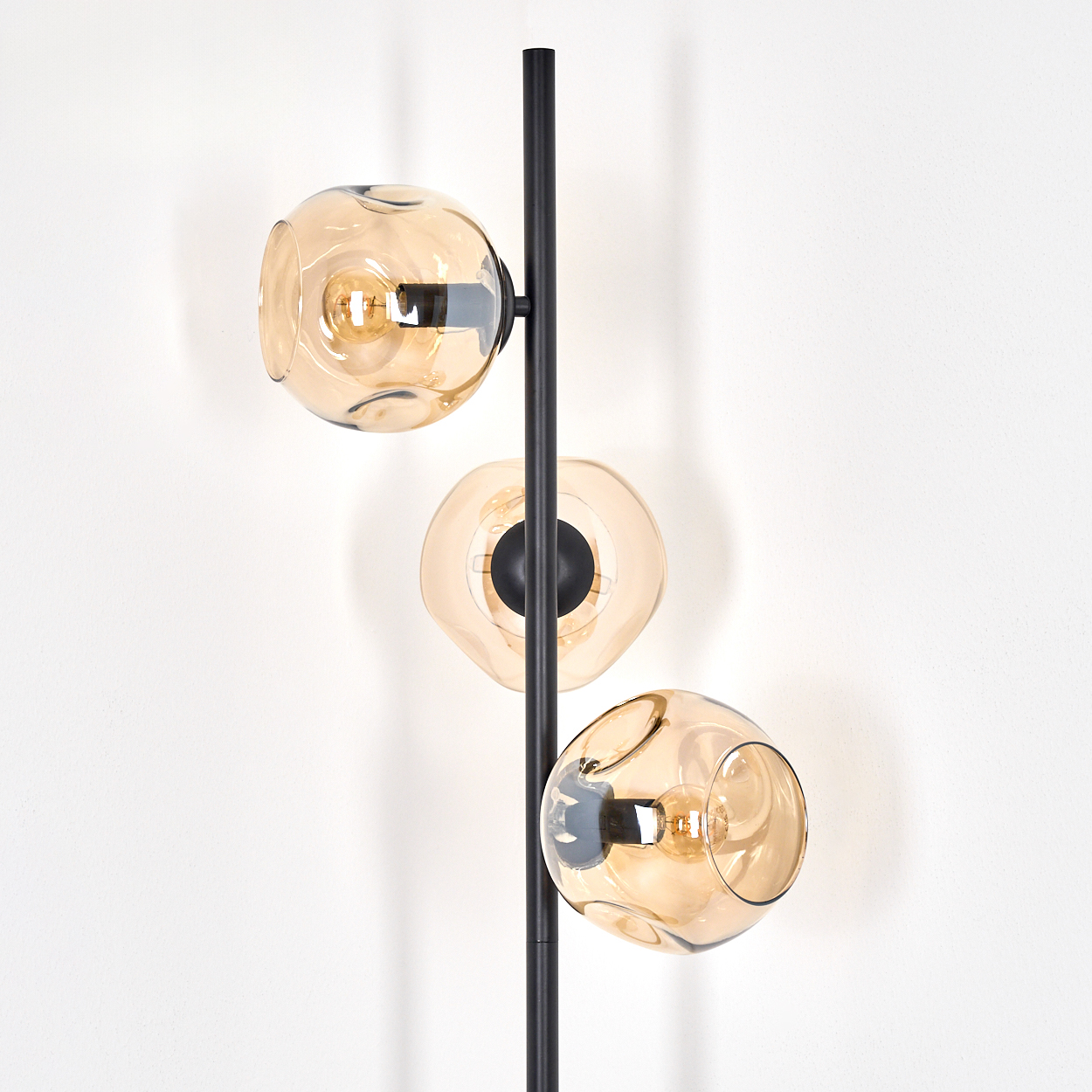 Eine moderne Stehlampe mit drei bernsteinfarbenen Glaskugeln, in denen sich jeweils eine Glühbirne befindet. Die Lampe hat ein schwarzes Metallgestell vor einer weißen Wand.
