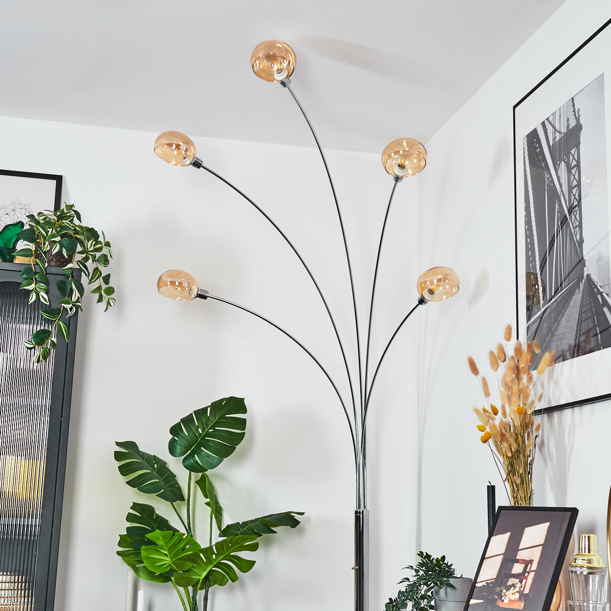 Eine moderne Stehlampe mit fünf bernsteinfarbenen Glühbirnen, umgeben von Pflanzen und gerahmten Kunstwerken. Der Raum hat weiße Wände.