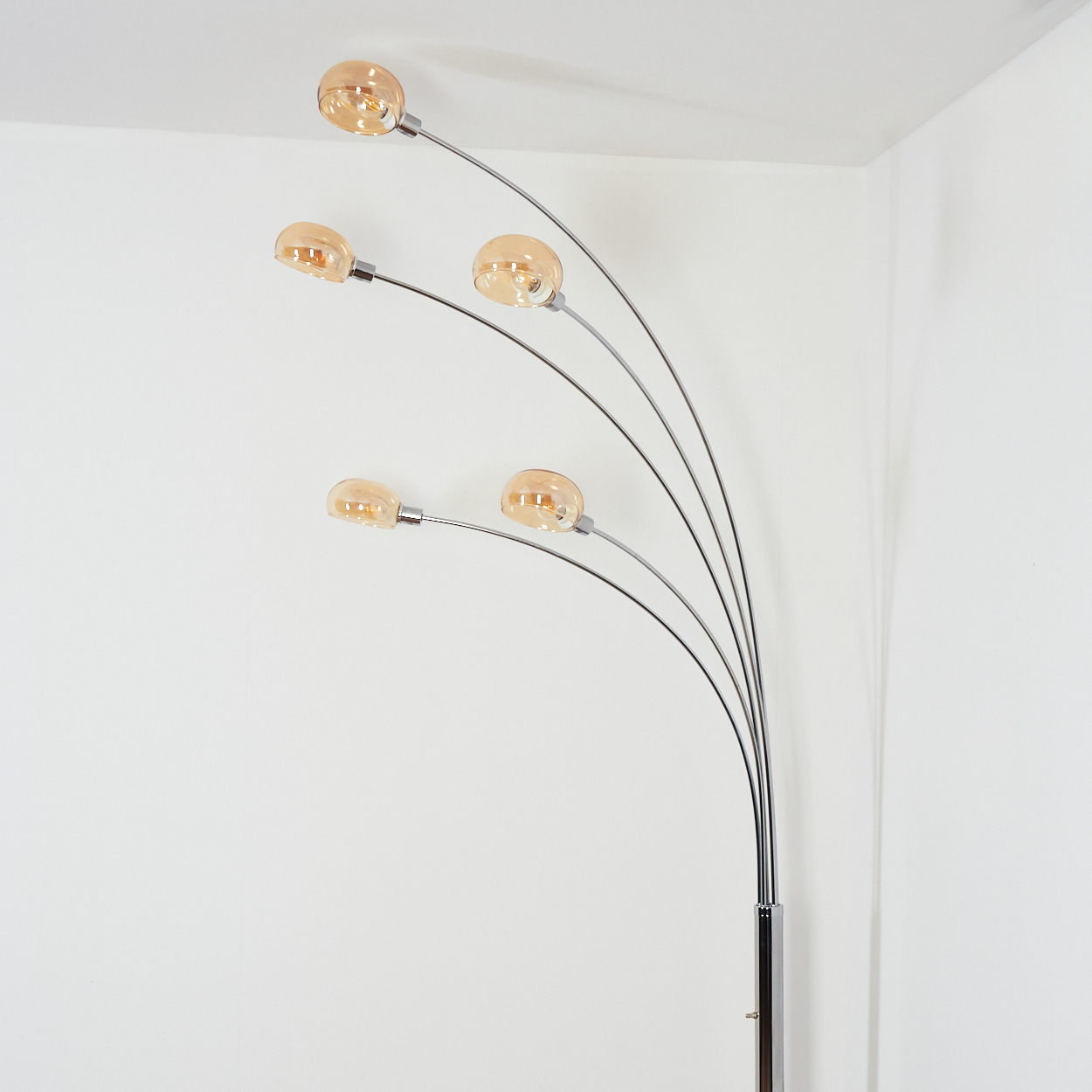 Eine moderne Bogenstehlampe mit fünf bernsteinfarbenen Kugelleuchten. Die Lampe hat eine Chromoberfläche und steht in einer weißen Ecke.