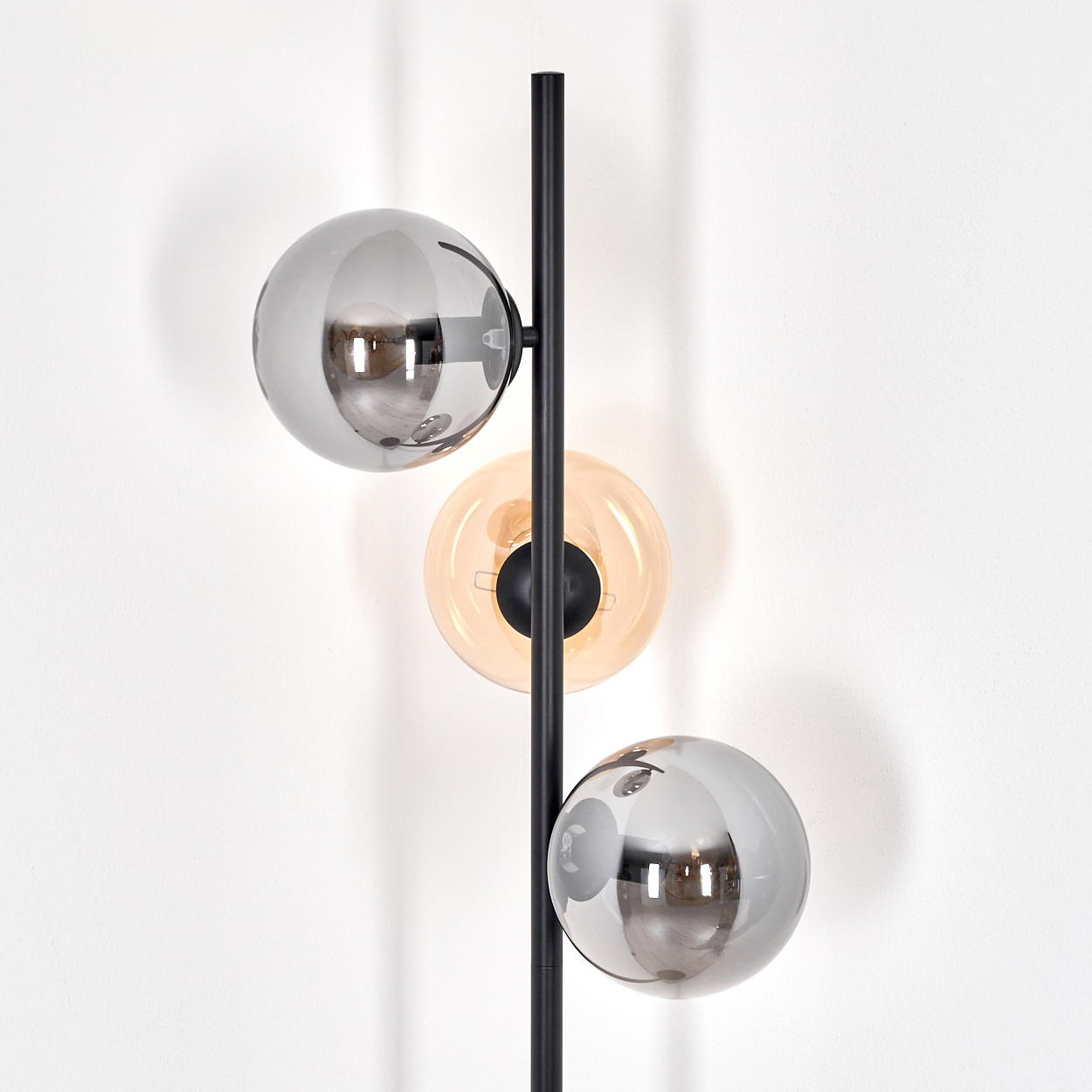 Eine moderne Stehlampe mit drei Glaskugeln in Grau und Bernstein vor einer weißen Wand. Die Lampe hat einen schwarzen Stab.