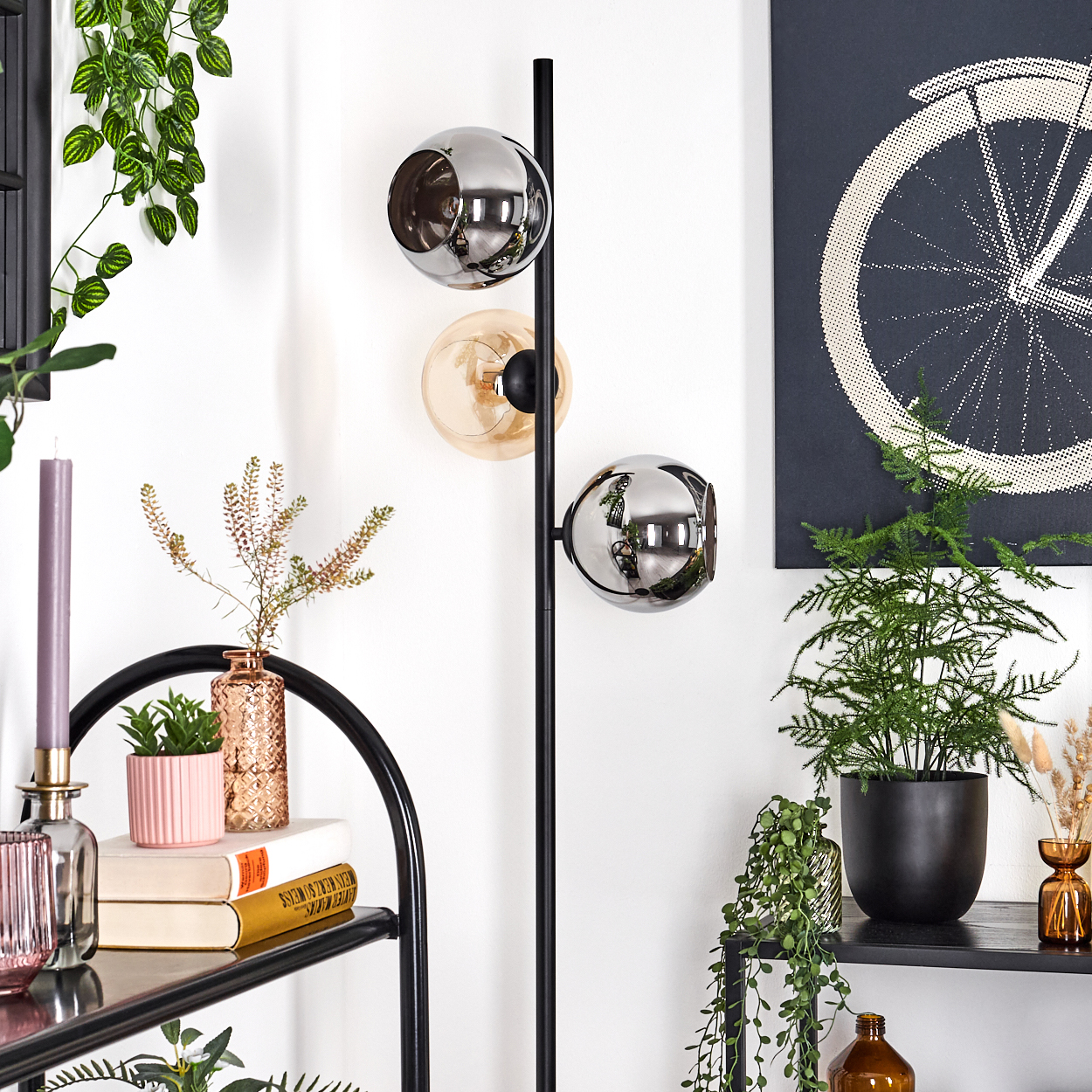 Ein modernes Interieur mit einer schwarzen Lampe mit drei kugelförmigen Lichtern. Es gibt Pflanzen, Bücher und Kunstwerke. Ein Fahrradrad ist an der Wand.