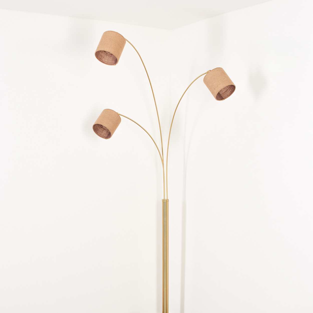 Eine Stehlampe mit drei Lampenschirmen. Die Lampenschirme sind beige zylindrisch, und der Ständer ist golden. Der Hintergrund ist eine weiße Wand.