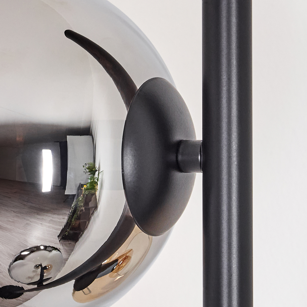 Nahaufnahme einer modernen Lampe mit reflektierendem Globus und schwarzen Metallelementen. Die Reflexion zeigt einen Raum mit Pflanzen und einem Fenster.