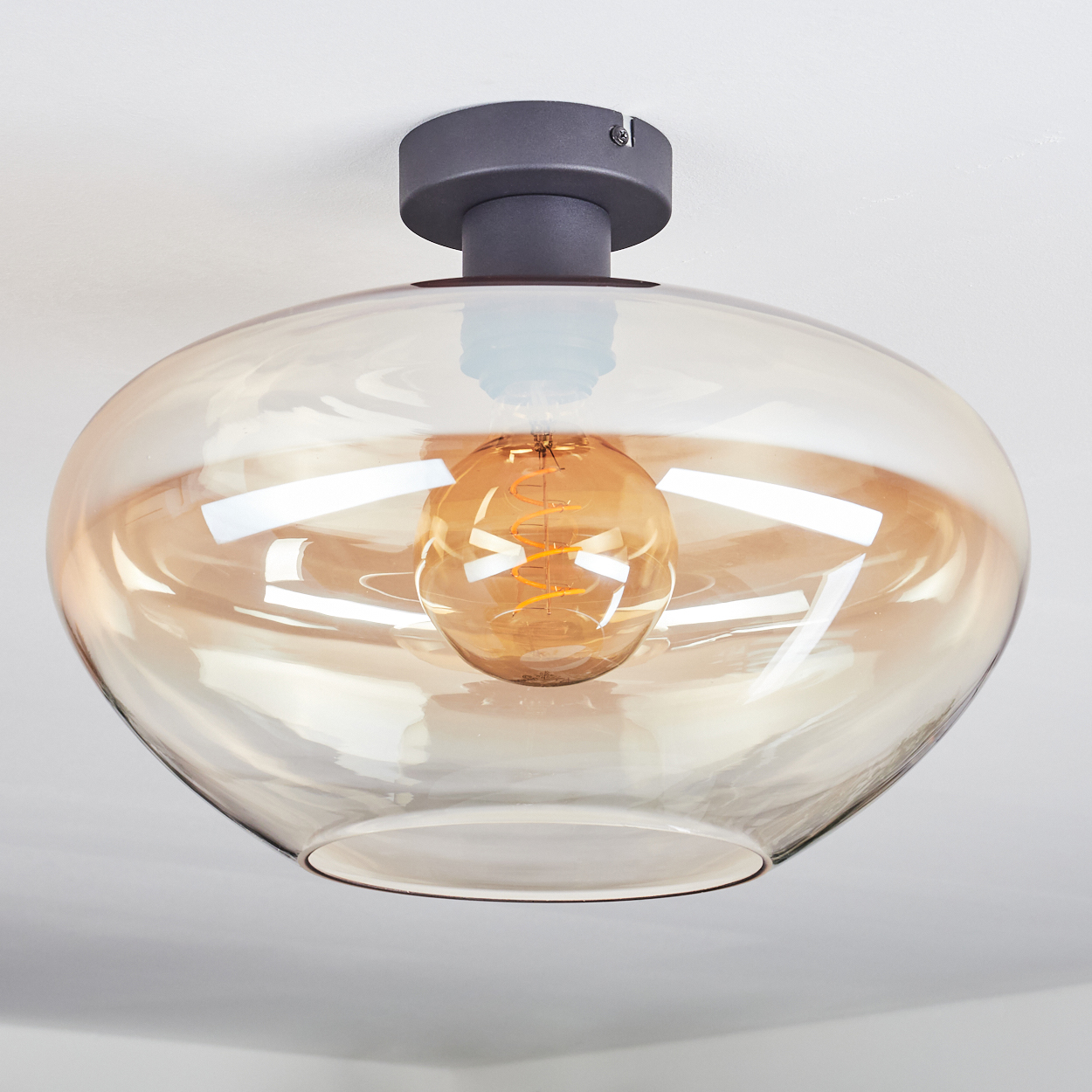 Eine moderne Deckenleuchte. Sie hat einen bronzefarbenen Glasschirm mit einer Glühbirne im Inneren. Der Sockel ist dunkelgrau, die Decke weiß.