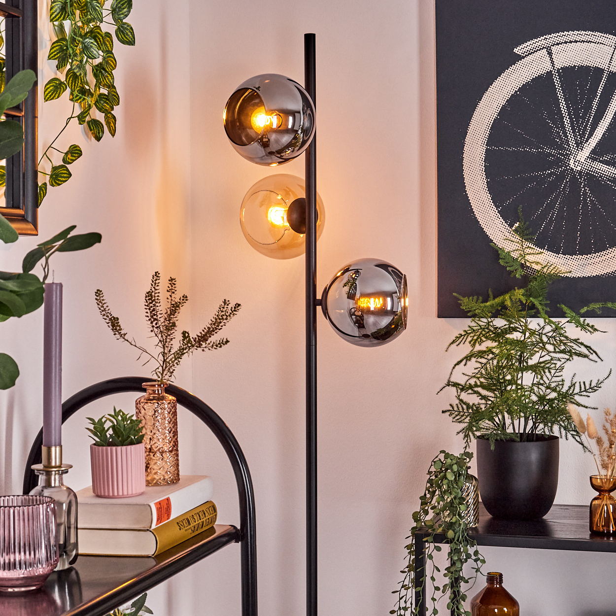 Eine moderne Lampe mit drei Glaskugeln beleuchtet einen Raum. Pflanzen, Bücher und Kunstwerke dekorieren den Raum und schaffen eine gemütliche Atmosphäre.