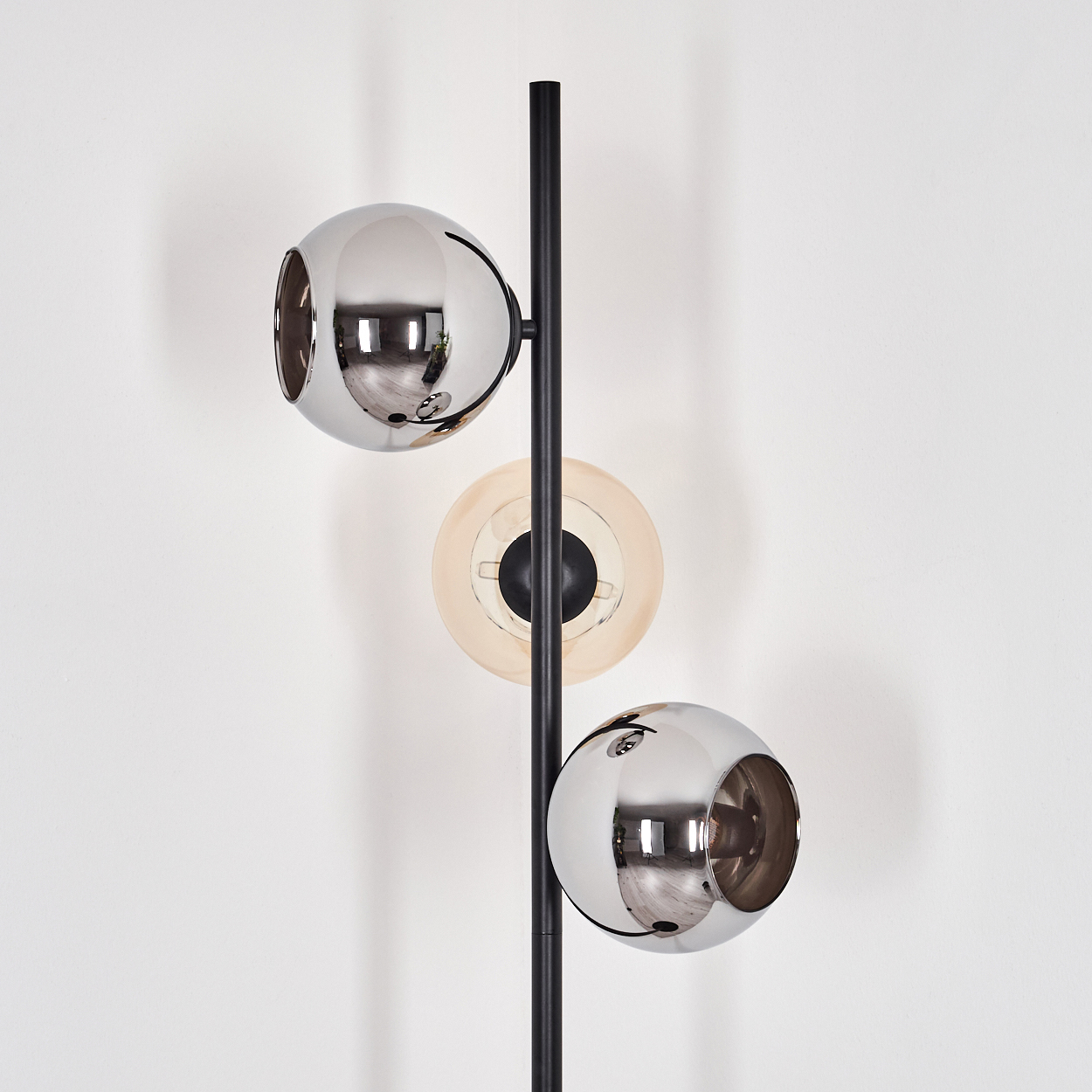Eine moderne Lampe mit einem schwarzen Pfahl und drei kugelförmigen Lichtern. Zwei sind verchromt, eines ist beige. Der Hintergrund ist eine weiße Wand.