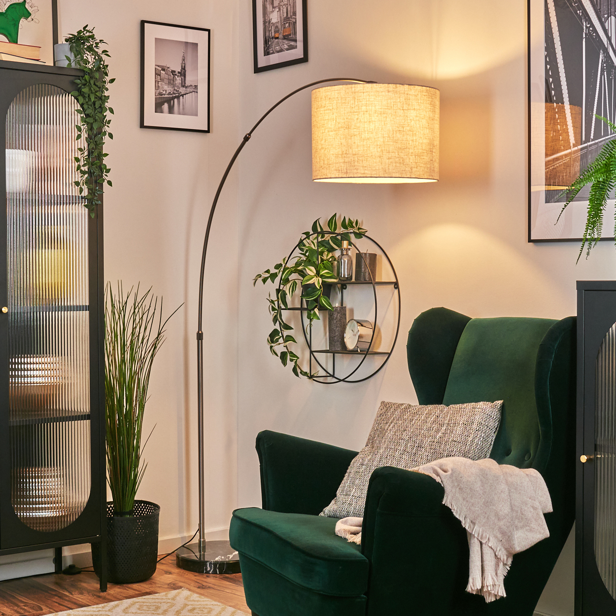 Ein grüner Sessel mit Kissen und einer Decke. Eine Lampe, ein Pflanzenregal, ein Schrank und gerahmte Bilder vervollständigen das gemütliche Interieur.