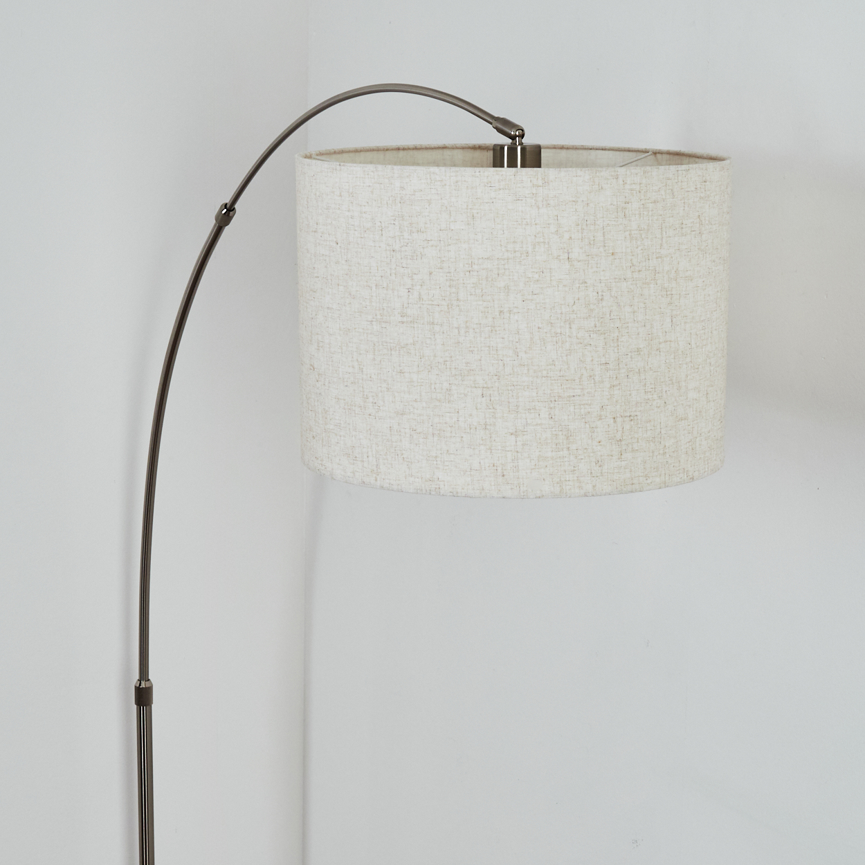 Eine moderne Stehlampe mit gebogenem, dunklem Metallrahmen. Sie hat einen beigefarbenen Stofflampenschirm vor einer weißen Wand.