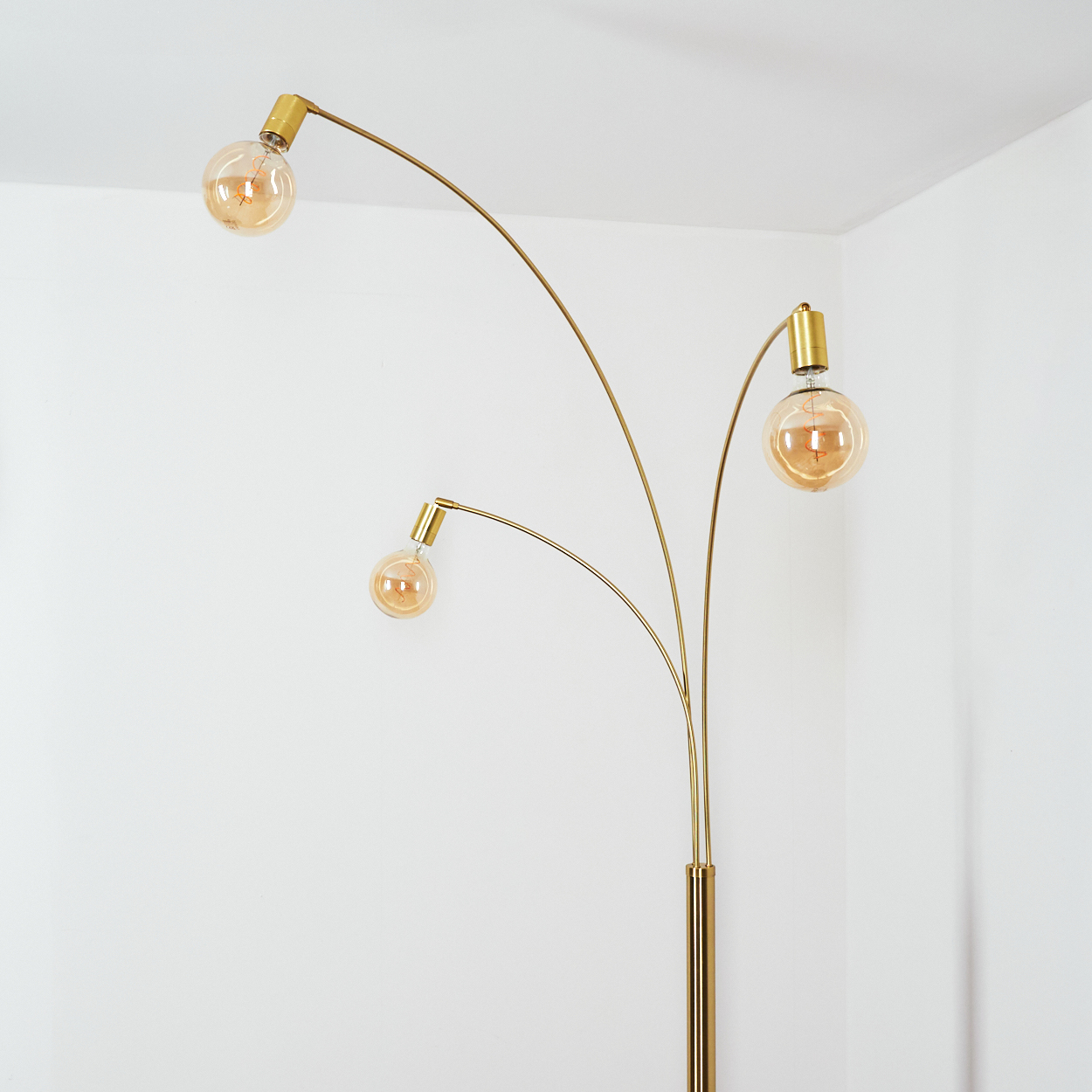 Eine goldene Stehlampe mit drei Glühbirnen. Die Glühbirnen sind rund und leuchten. Der Hintergrund ist eine weiße Wand in einer Ecke.