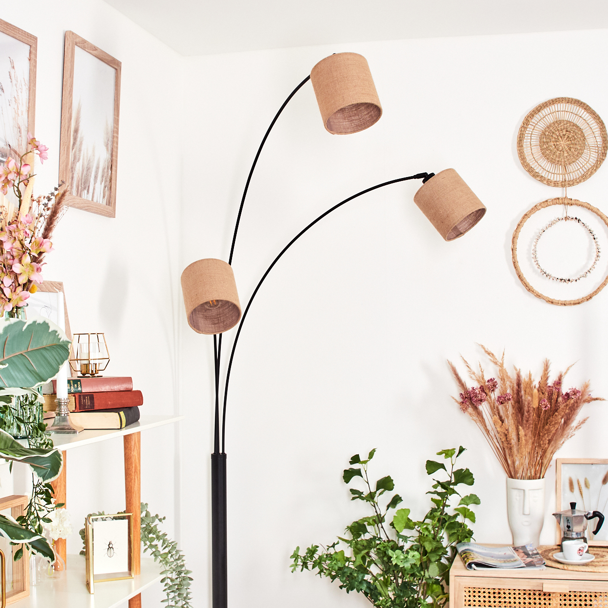 Ein modernes Wohnzimmer mit Stehlampe, Pflanzen und dekorativen Gegenständen. Die Lampe hat drei beige Schirme. Weiße Wände.