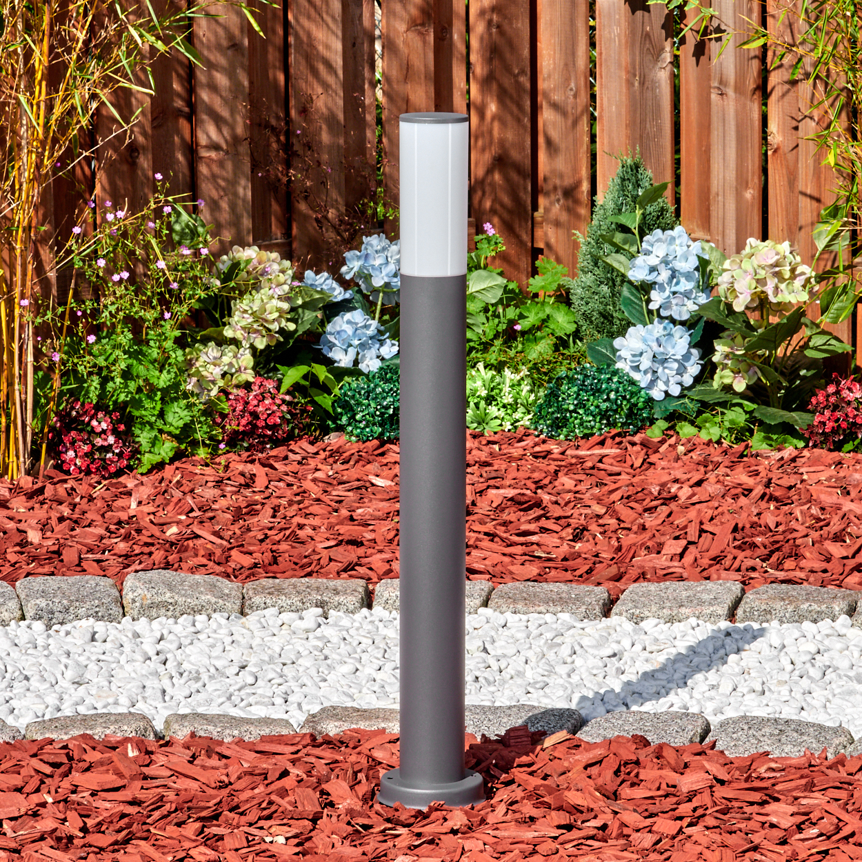 Eine graue Außenlampe steht in einem Gartenbeet. Das Beet hat rote Mulch, weiße Steine und bunte Blumen. Im Hintergrund ist ein Holzzaun zu sehen.