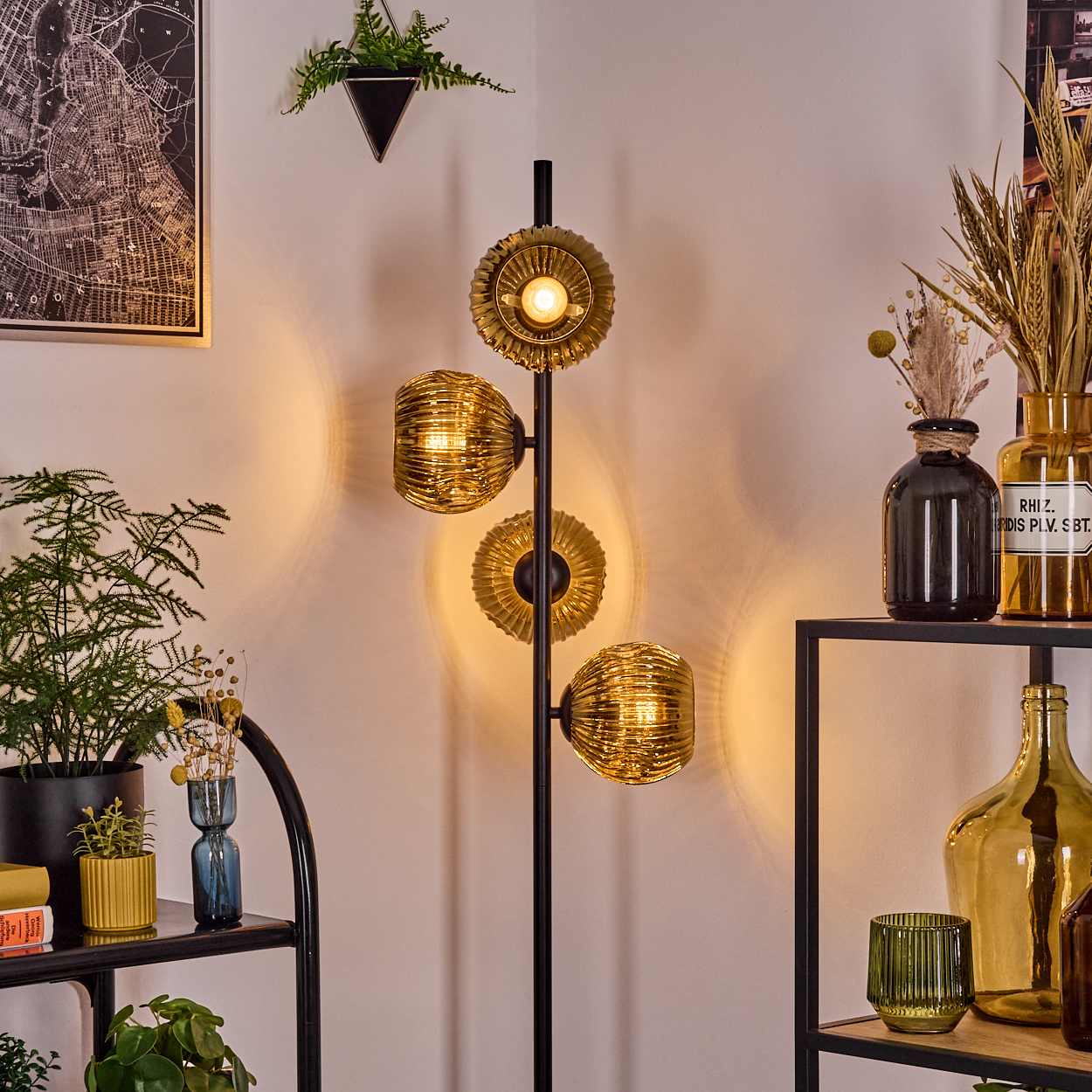 Stehlampe mit drei goldenen Glasschirmen. Pflanzen und Vasen stehen auf Regalen. Eine Karte hängt an der Wand.