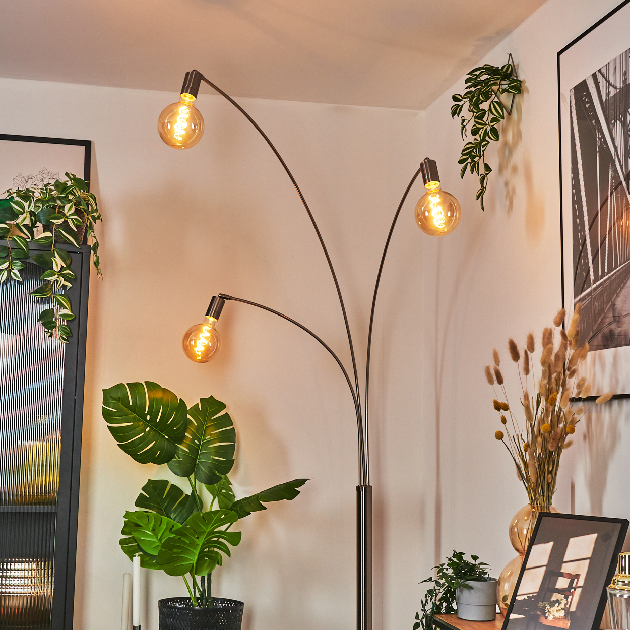Eine moderne Stehlampe mit drei Glühbirnen beleuchtet einen Raum mit Pflanzen und Dekoration. Ein gerahmtes Brückenfoto hängt an der Wand.