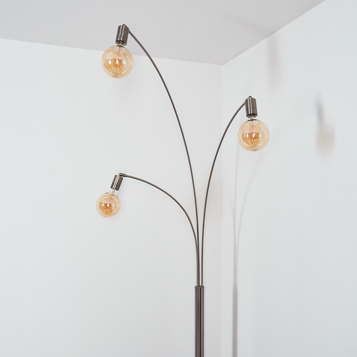 Eine moderne Stehlampe mit drei kugelförmigen Glühbirnen. Das Gestell ist metallgrau, vor einer weißen Wand.