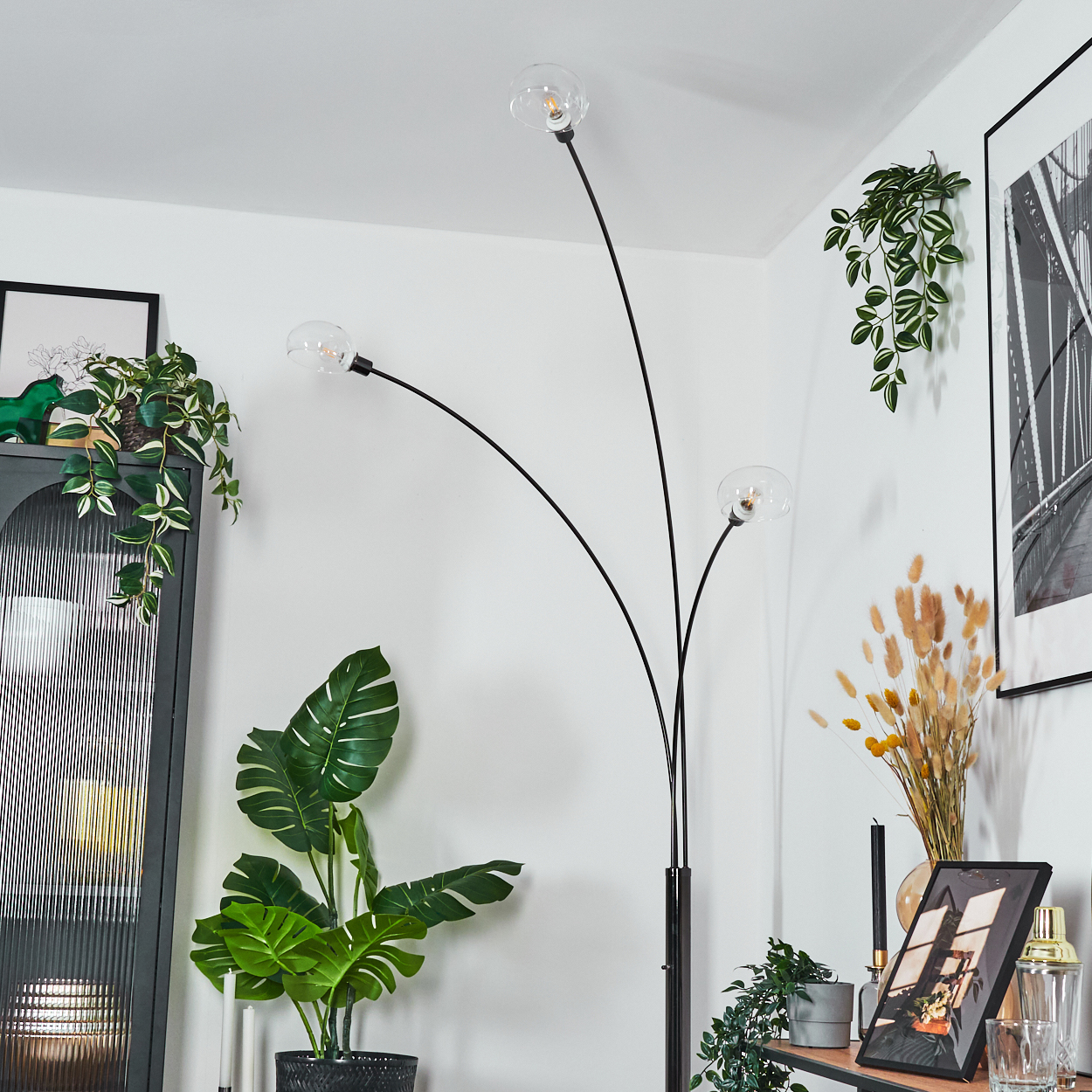 Ein modernes Wohnzimmer mit schwarzer Stehlampe, Pflanzen, einem gerahmten Bild und Dekorationsartikeln. Die Wände sind weiß.