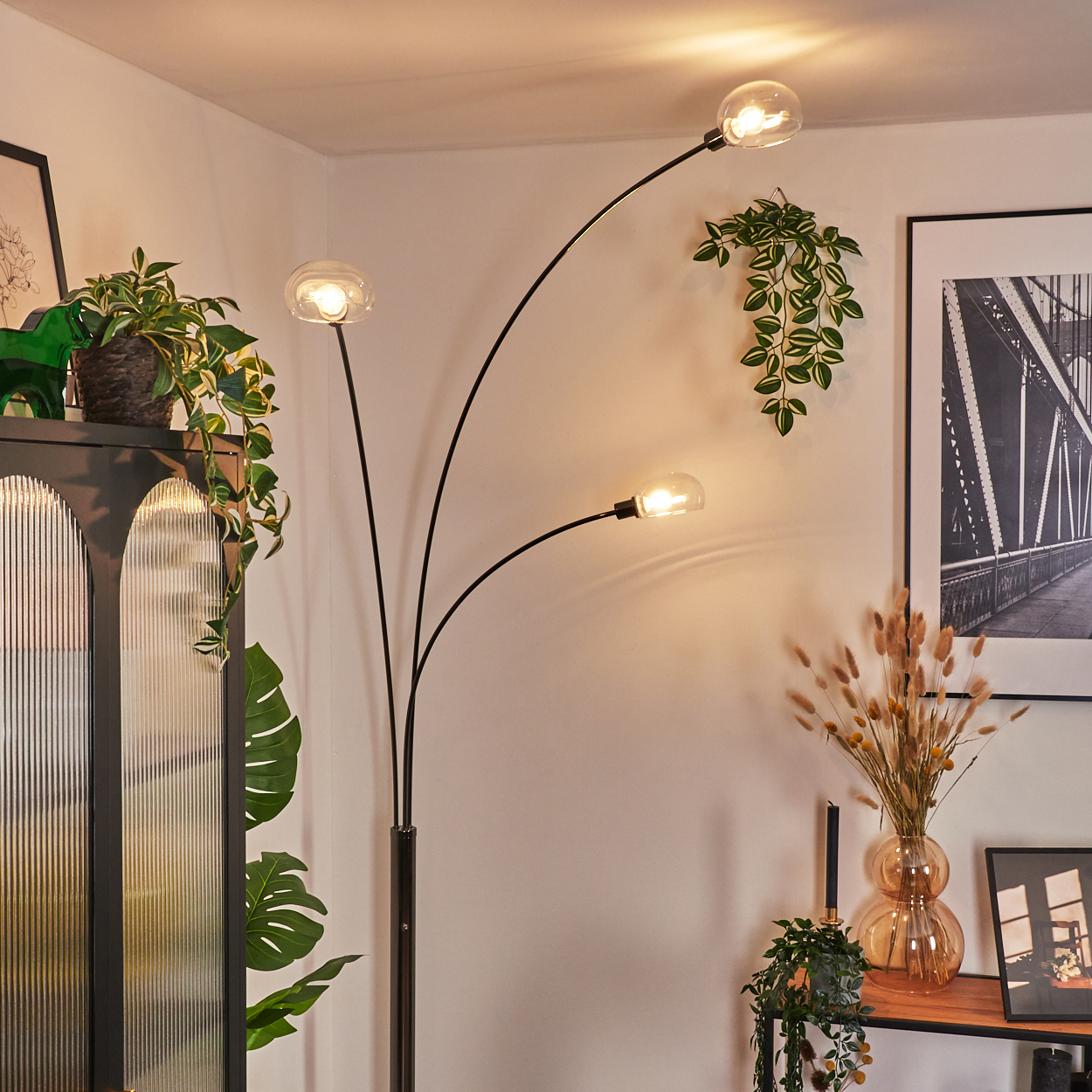 Eine moderne Stehlampe mit drei Glühbirnen beleuchtet einen Raum mit Pflanzen, einem Schrank und einem Brückenbild.