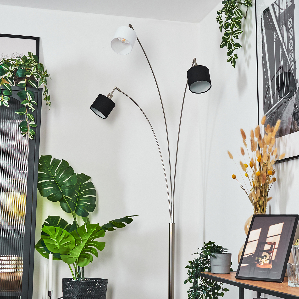 Ein modernes Wohnzimmer mit einer Stehlampe mit drei Köpfen, einer Monstera-Pflanze, gerahmten Bildern und dekorativen Pflanzen.