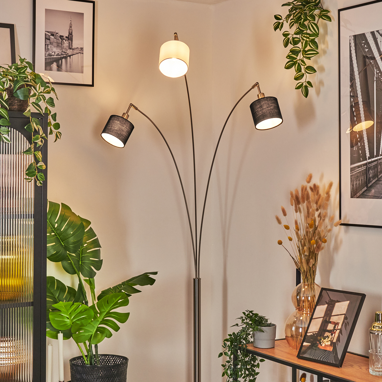Eine Stehlampe mit drei Lichtern. Pflanzen in Töpfen und gerahmte Bilder im Hintergrund. Die Wand ist beige.