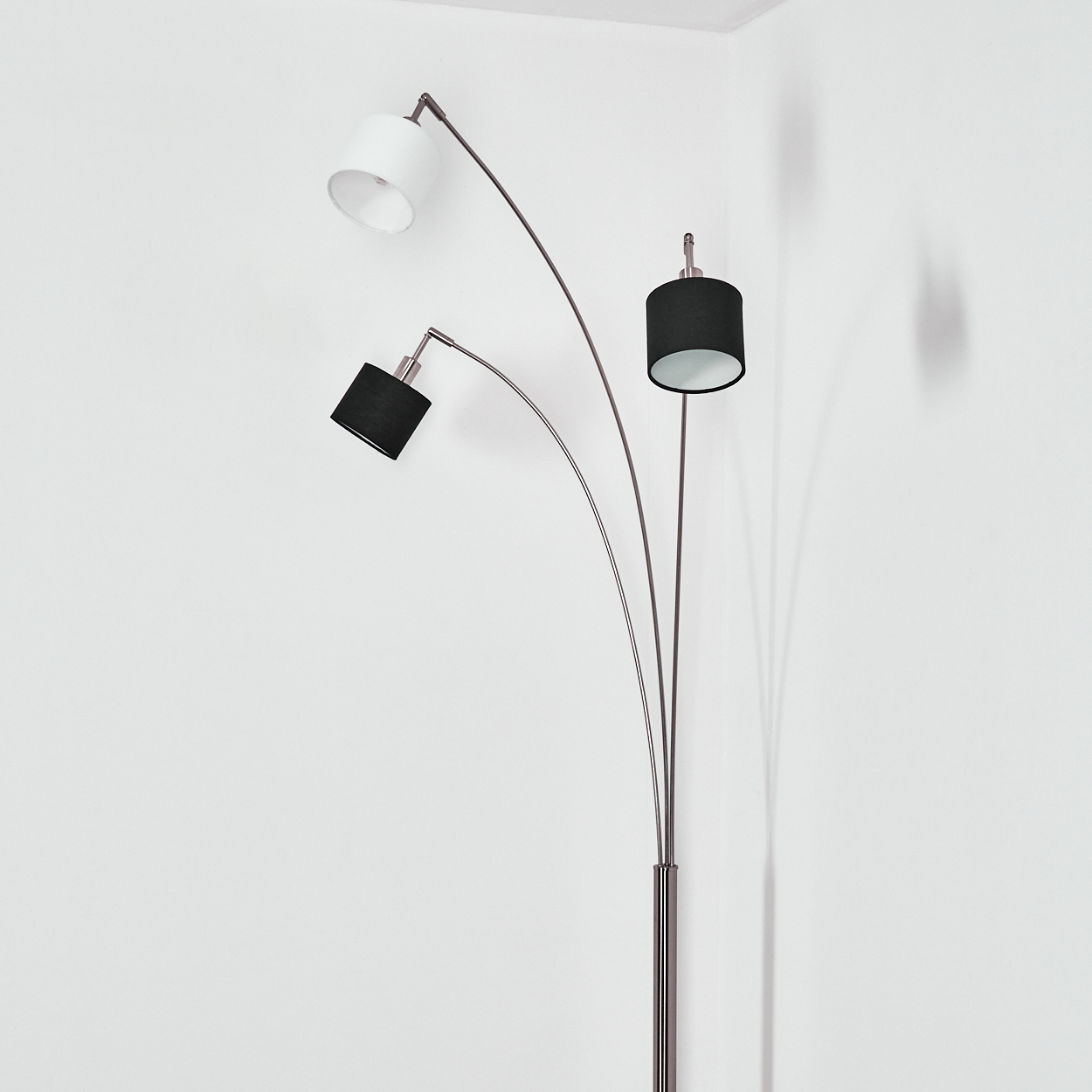 Eine moderne Stehlampe mit drei Lampenschirmen. Einer ist weiß, zwei sind schwarz. Die Lampe hat ein silbernes Metallgestell. Sie steht in einer Ecke.