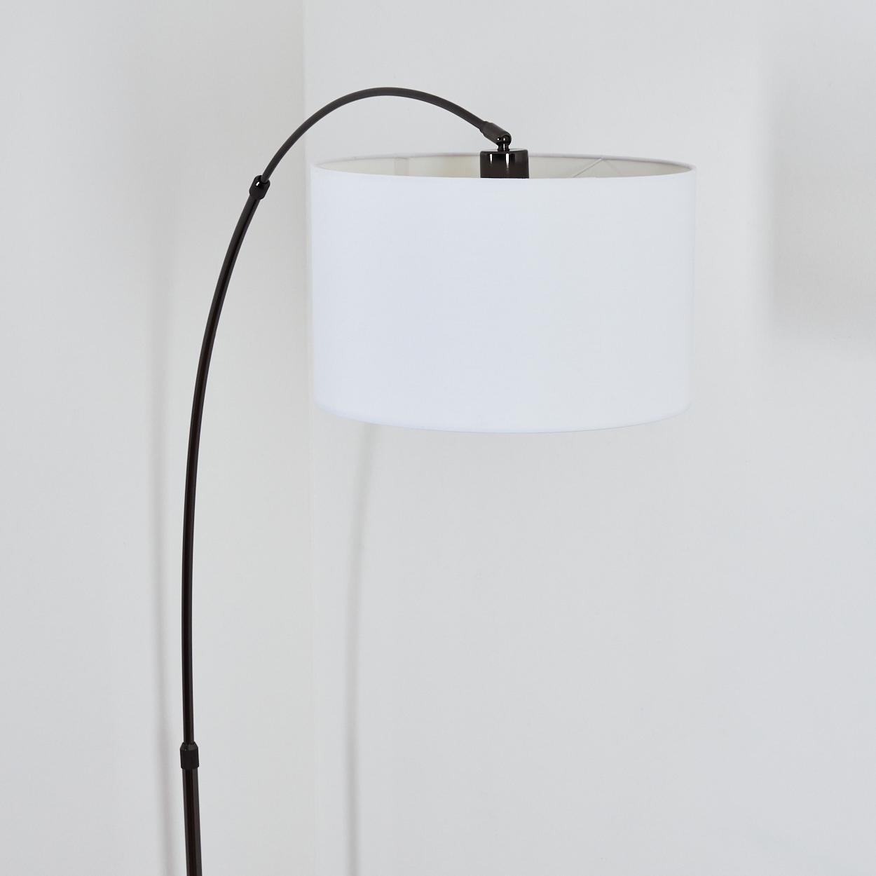 Eine Stehlampe mit weißem Lampenschirm und dunklem Metallrahmen. Sie steht in einer Ecke vor einer weißen Wand.