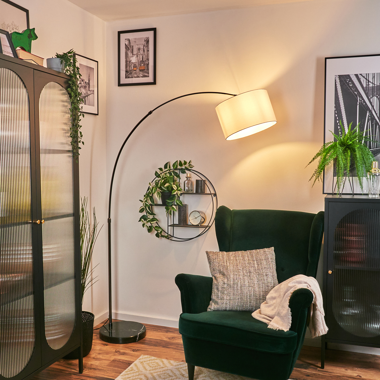 Ein grüner Sessel mit Kissen und Decke. Eine Stehlampe beleuchtet den Raum. Es gibt Pflanzen, Bilder und einen Schrank.