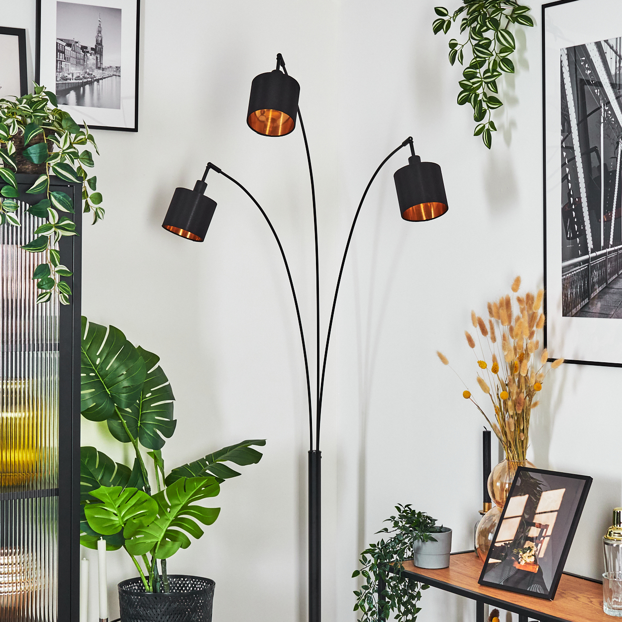 Eine schwarze Stehlampe mit drei Lampenschirmen. Pflanzen und Bilder sind im Hintergrund.