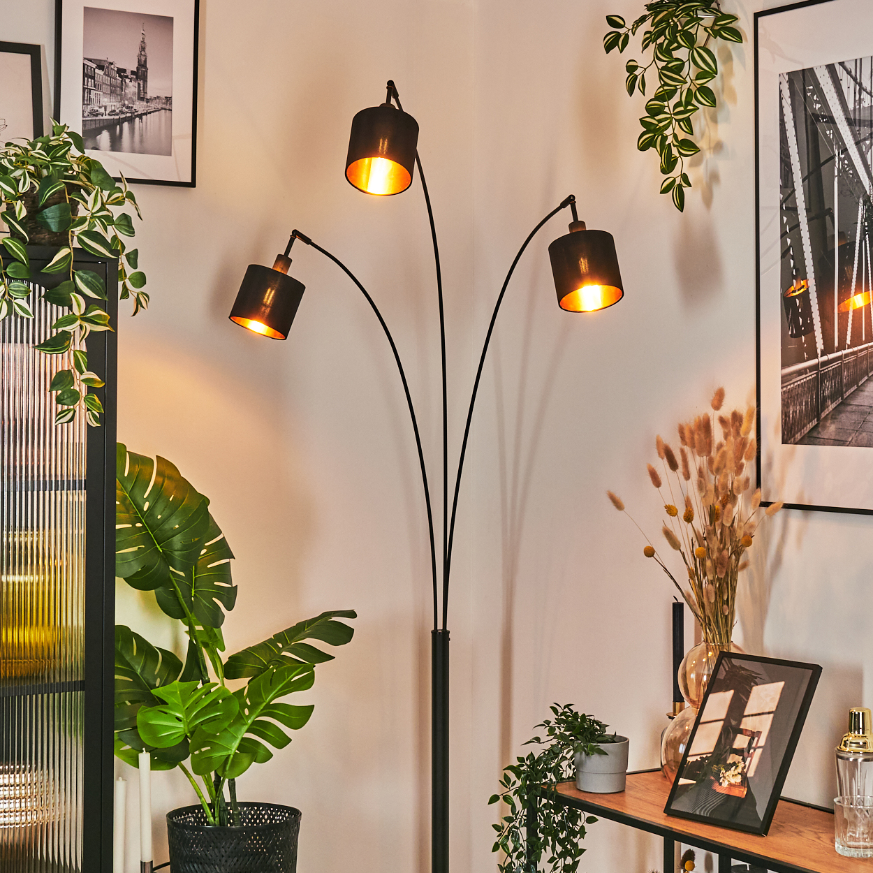 Eine moderne Stehlampe mit drei Lichtern steht in einer Ecke. Pflanzen und gerahmte Bilder sind sichtbar und schaffen eine gemütliche Atmosphäre.