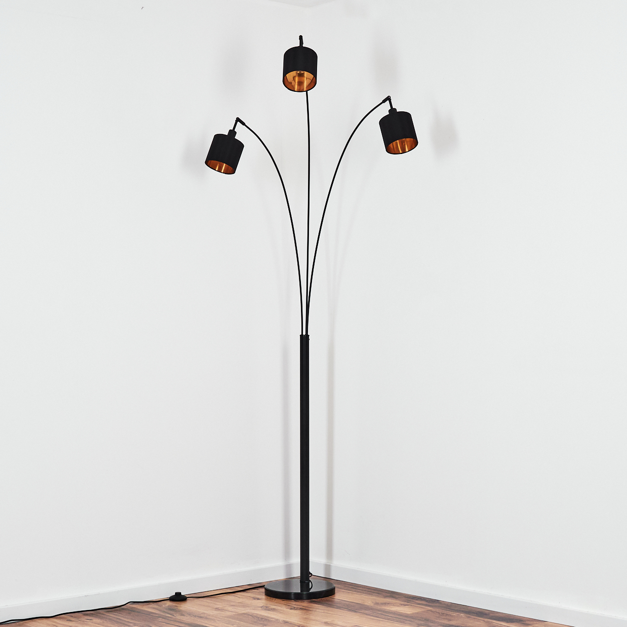 Eine schwarze Stehlampe mit drei Lichtern. Die Schirme sind schwarz mit goldenem Innenraum. Sie steht in der Ecke eines Raumes mit Holzböden.