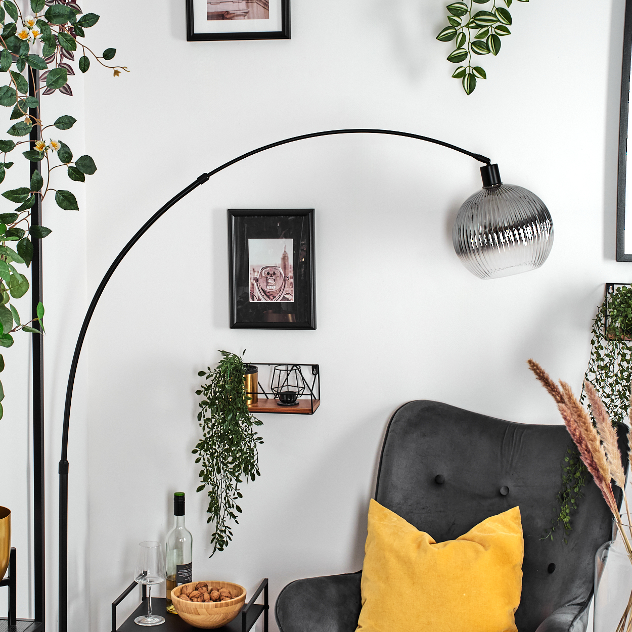 Wohnzimmerszene mit schwarzer Bogenlampe, grauem Sessel mit gelbem Kissen und Zierpflanzen. Bilder an einer weißen Wand.