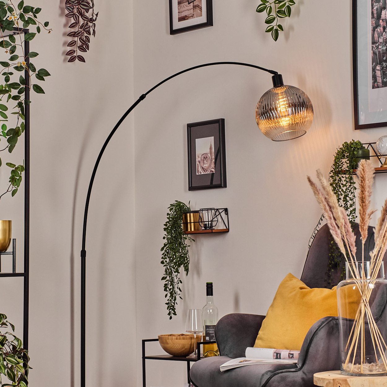 Modernes Wohnzimmer mit schwarzer Bogenlampe, grauem Sessel, gelbem Kissen und Zierpflanzen. Eine Glasvase mit getrockneten Blumen.