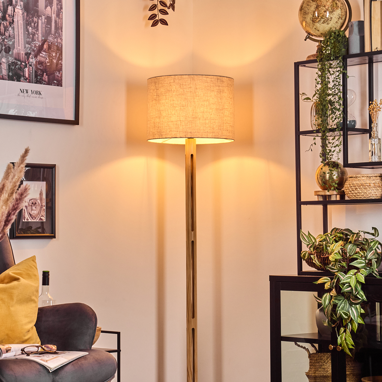 Eine Stehlampe steht in einem Raum mit einem Regal voller Pflanzen, einem Globus und gerahmter Kunst. Ein Sessel mit gelbem Kissen ist sichtbar.