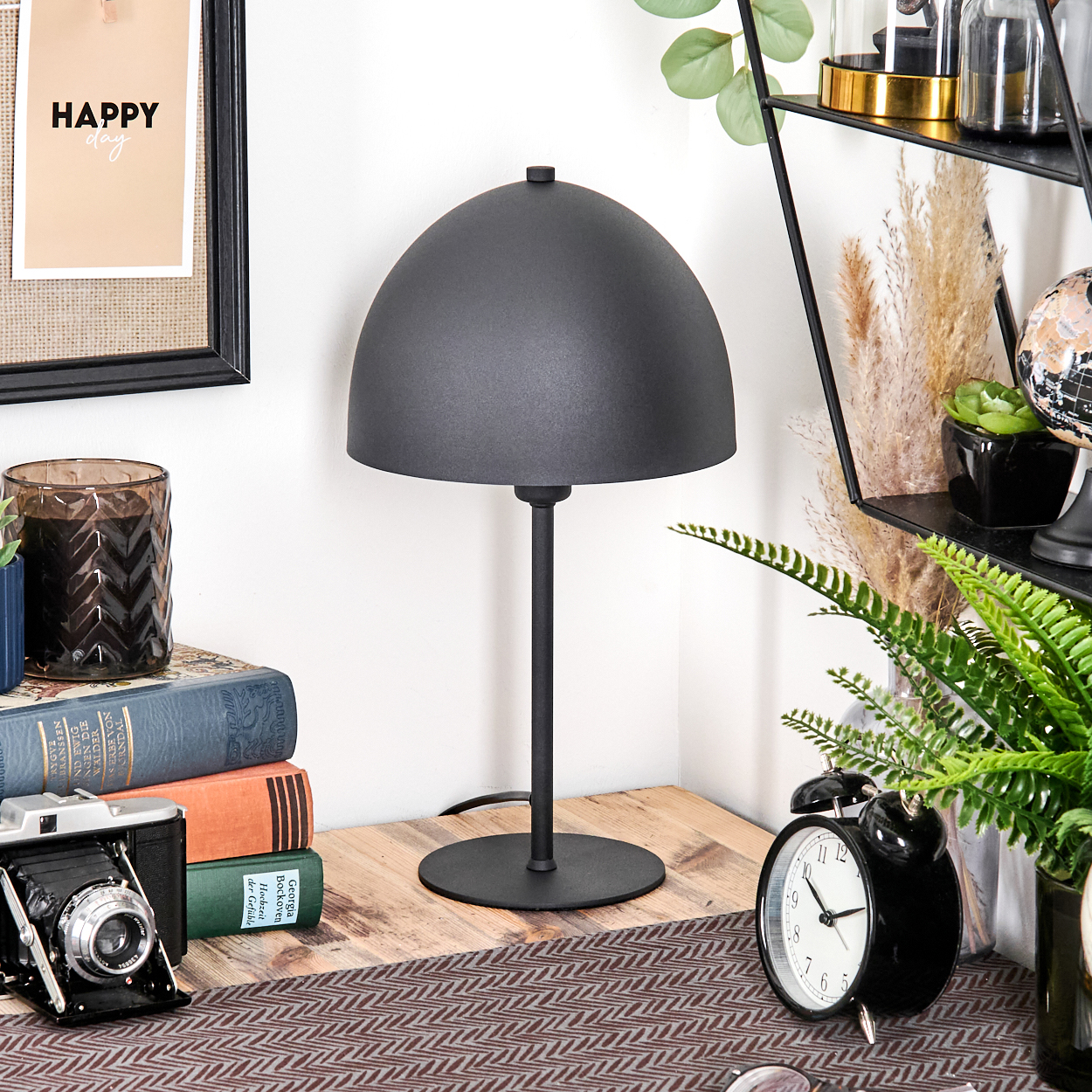 Eine schwarze Lampe, Bücher, eine Kamera, eine Uhr und Pflanzen stehen auf einem Holztisch. Ein Bild mit 'Happy day' an der Wand.