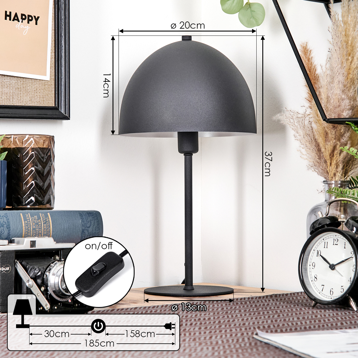 Eine schwarze Schreibtischlampe mit rundem Lampenschirm, schwarzem Fuß und Schalter. Ein Wecker, Bücher und ein Rahmen sind sichtbar.