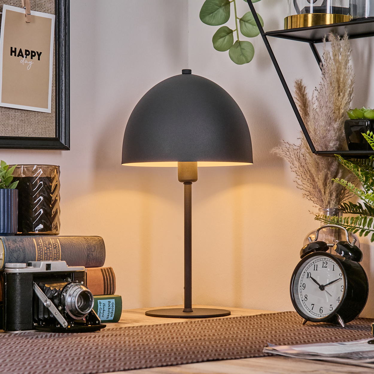 Eine Schreibtischlampe mit schwarzem Schirm steht auf einer Holzoberfläche. Eine alte Kamera, Bücher und eine Uhr sind sichtbar. Eine fröhliche Notiz hängt an der Wand.