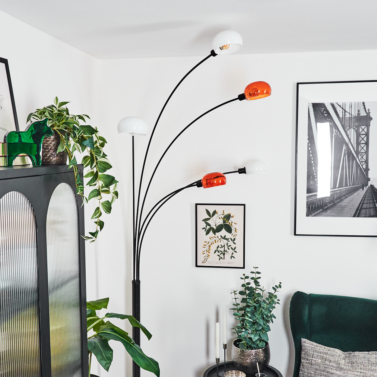 Ein modernes Wohnzimmer mit hoher Lampe, Pflanzen, dunklem Schrank und gerahmtem Brückenfoto. Rechts ein grüner Sessel.