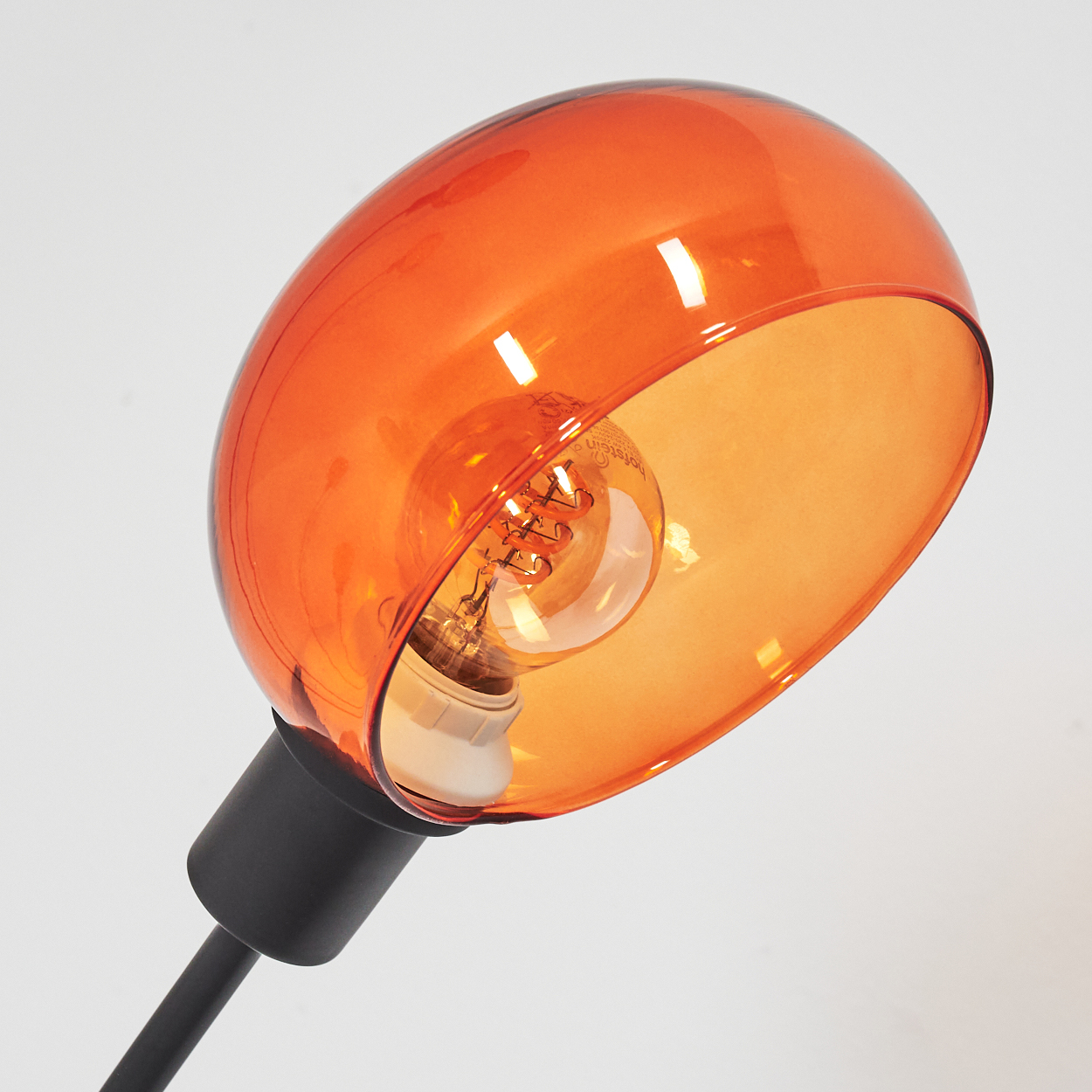 Nahaufnahme einer modernen Lampe. Sie hat einen orangefarbenen Lampenschirm, eine Glühbirne und einen schwarzen Ständer. Der Hintergrund ist weiß.