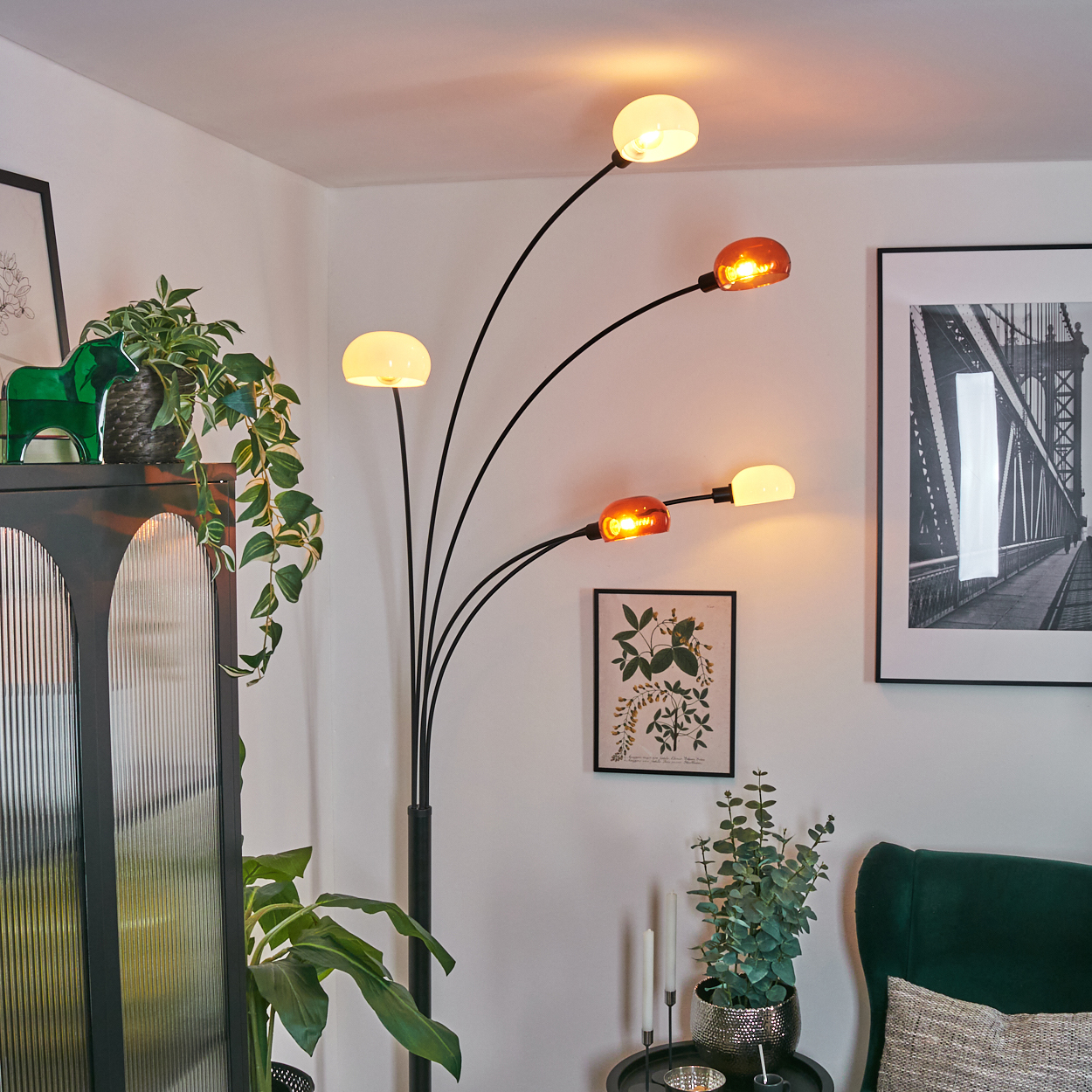 Ein modernes Wohnzimmer mit einer schwarzen Stehlampe mit mehreren Lichtern. Es gibt einen Schrank, Pflanzen, gerahmte Kunst und einen grünen Sessel.