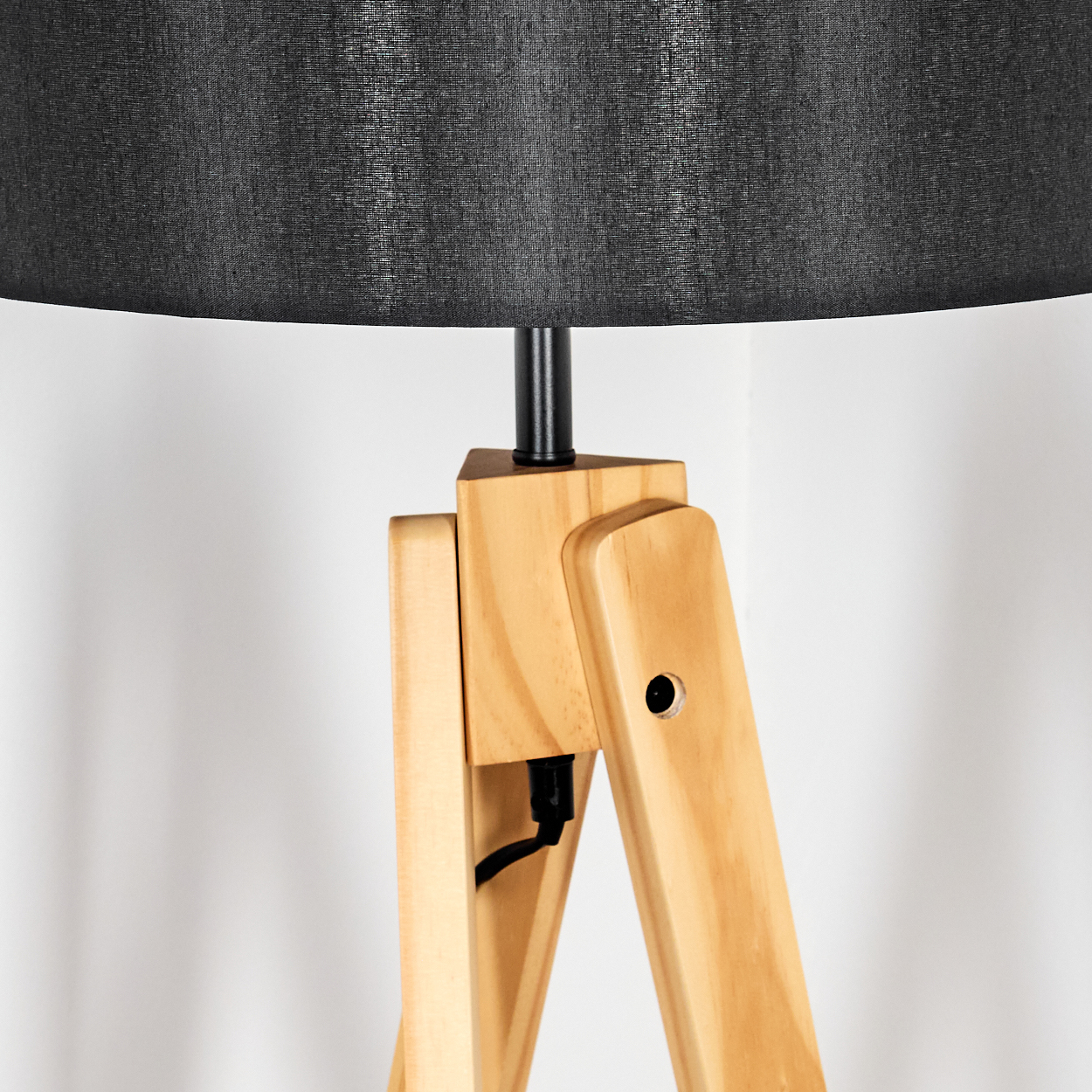 Nahaufnahme einer hölzernen Stativlampe mit schwarzem Lampenschirm. Die Holzbeine sind hellbraun, der Hintergrund ist weiß.