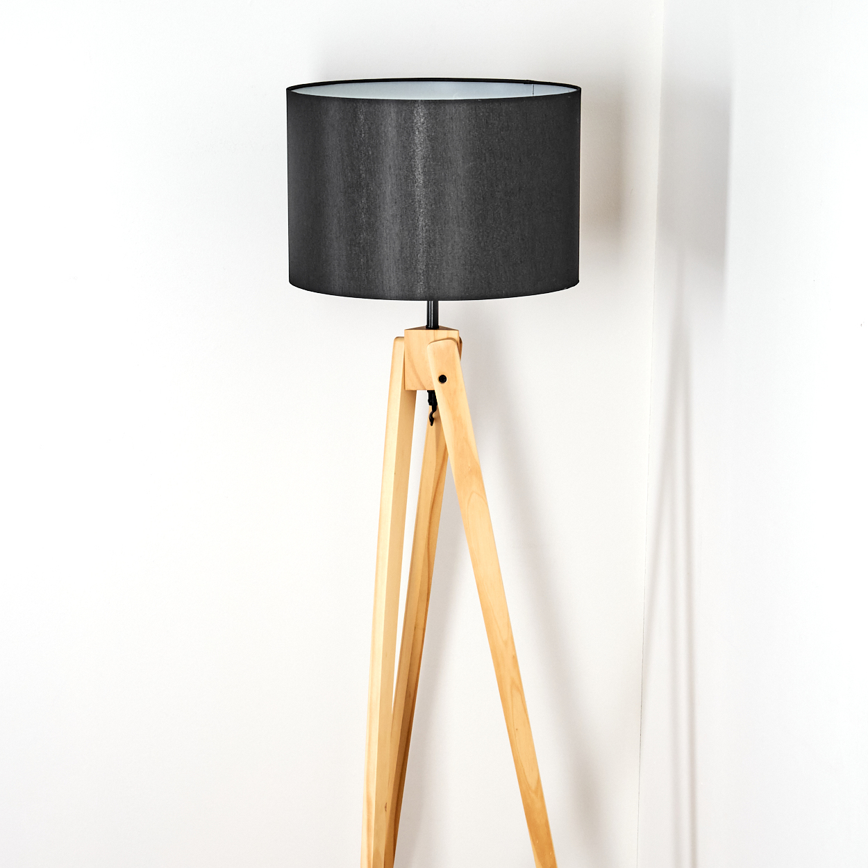 Eine moderne Stehlampe aus Holz mit schwarzem Lampenschirm steht vor einer weißen Wand. Die Lampe hat ein Stativ.