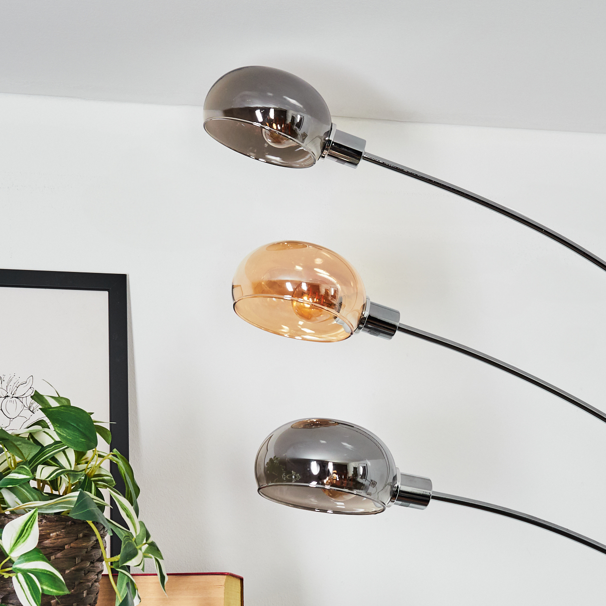 Eine moderne Stehlampe mit drei Chromarmen und Glasschirmen. Einer ist bernsteinfarben, zwei sind grau. Links sind eine Pflanze und ein Bilderrahmen.