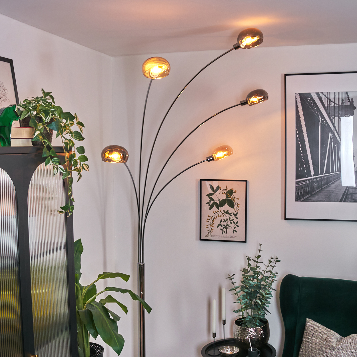 Ein modernes Wohnzimmer mit einer schwarzen Stehlampe mit fünf Glühbirnen. Pflanzen und gerahmte Kunstwerke schmücken die weißen Wände. Ein grüner Sessel.