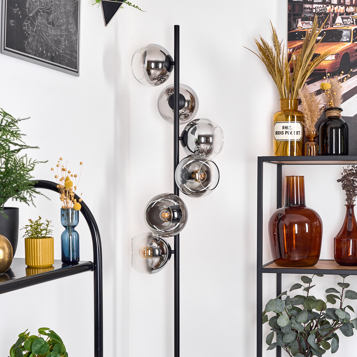 Eine moderne Stehlampe mit sechs Glaskugeln. Regale mit Vasen und Pflanzen befinden sich im Hintergrund, zusammen mit einer Karte und einem Poster.