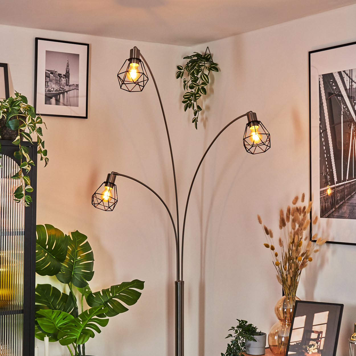 Ein modernes Wohnzimmer mit Stehlampe, Pflanzen und gerahmten Bildern. Die Lampe hat drei Lichter. Es gibt eine Vase mit Trockenblumen.