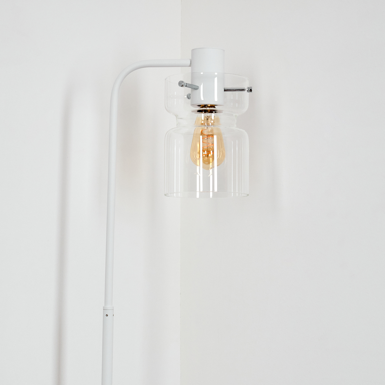 Eine weiße, moderne Stehlampe mit Glasschirm und sichtbarer Edison-Glühbirne vor weißer Wand.