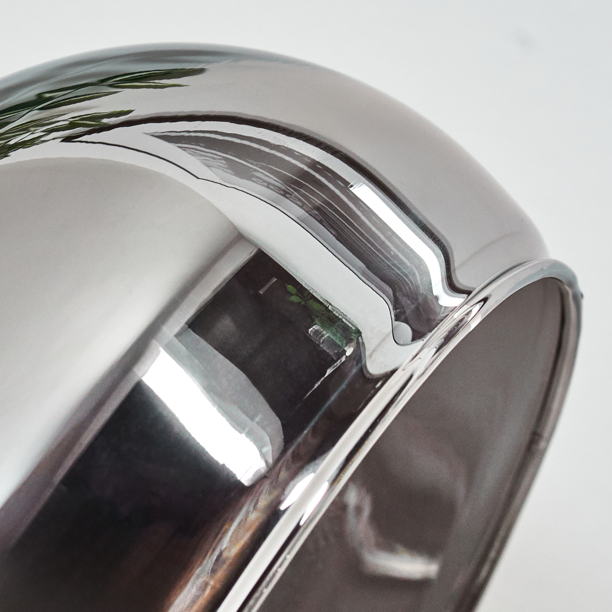 Nahaufnahme eines glänzenden, metallischen Objekts mit gebogener Form, das eine grüne Pflanze und einen weißen Hintergrund reflektiert.